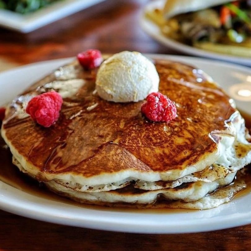 Stack of golden-brown pancakes with syrup, a scoop of butter, and fresh raspberries on a white plate.