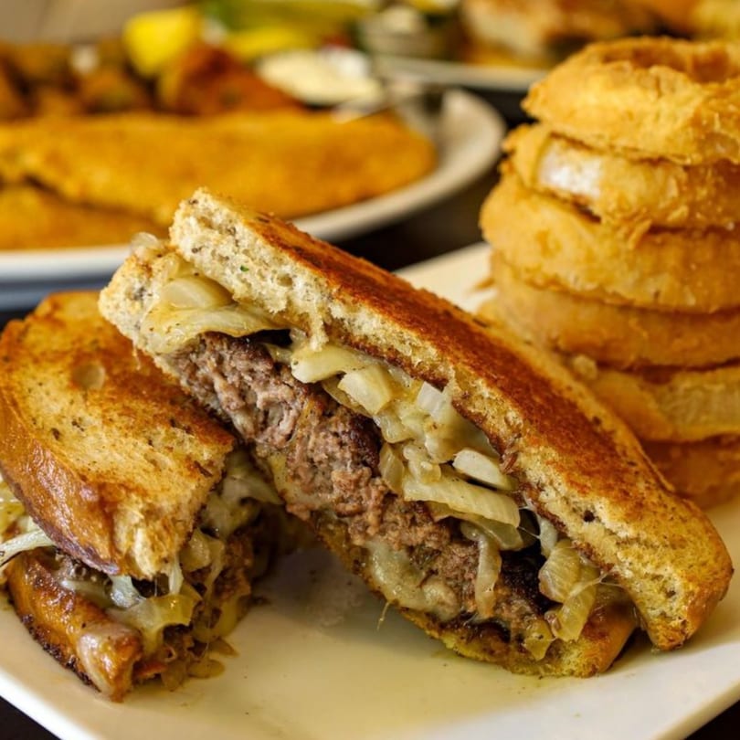 A grilled patty melt sandwich with caramelized onions and melted cheese, served with onion rings.